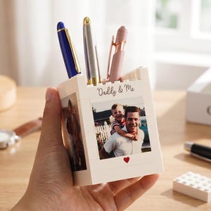 Può includere: Un portapenne bianco con una foto di un padre e un bambino e la scritta "Daddy & Me". Il portapenne contiene una penna blu, argento e rosa. Un orologio e una penna nera sono sulla superficie di legno.