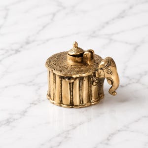 May include: A round brass container with a hinged lid featuring an intricate etched pattern on the top surface. The sides of the container have vertical ribbed detailing, and an elephant head sculpture is attached to the side as a decorative element.