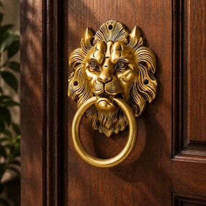 May include: A brass-colored metal door knocker shaped like a lion's head with a circular ring held in its mouth. The lion features detailed mane textures and facial features, mounted onto a dark brown wooden door with visible vertical grain.