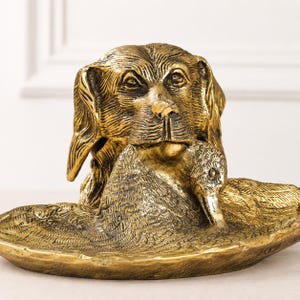 May include: A metallic gold-toned brass sculpture featuring a detailed dog head holding a duck in its mouth. The sculpture is integrated into a shallow, oval-shaped tray base with textured feather details across the surface of the duck.