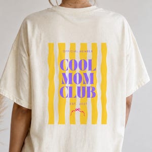Könnte beinhalten: Ein cremefarbenes Kurzarm-T-Shirt mit einem Rückendruck aus vertikalen, gelben, wellenförmigen Streifen und violettem Text, der OFFICIAL MEMBER COOL MOM CLUB EST. 2027 lautet, ergänzt durch eine kleine rote Schleifenillustration am unteren Rand.