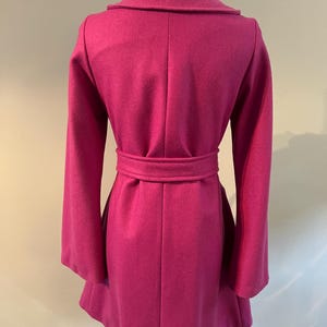 May include: A magenta coat on a mannequin, viewed from the back. It has a wide collar, long sleeves, and a belt at the waist. The coat's design includes a flared skirt and a tailored fit, showcasing a classic silhouette. The coat is made of a soft material.