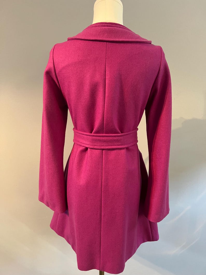 May include: A magenta coat on a mannequin, viewed from the back. It has a wide collar, long sleeves, and a belt at the waist. The coat's design includes a flared skirt and a tailored fit, showcasing a classic silhouette. The coat is made of a soft material.