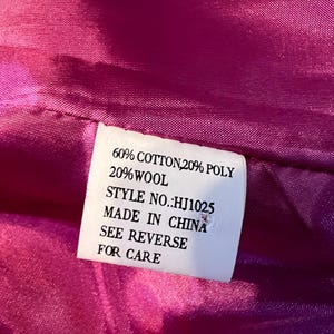 May include: Close-up of a pink fabric with a white label. The label states: 60% COTTON, 20% POLY, 20% WOOL, STYLE NO.: HJ1025, MADE IN CHINA, SEE REVERSE FOR CARE. The fabric has a glossy texture.