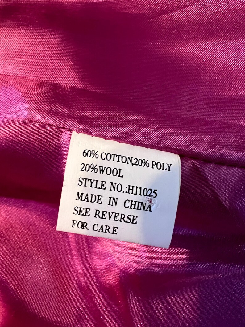 May include: Close-up of a pink fabric with a white label. The label states: 60% COTTON, 20% POLY, 20% WOOL, STYLE NO.: HJ1025, MADE IN CHINA, SEE REVERSE FOR CARE. The fabric has a glossy texture.