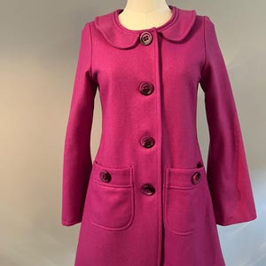 May include: A magenta coat with a rounded collar and large, matching buttons. The coat has two front pockets and an A-line shape. The coat is displayed on a mannequin.