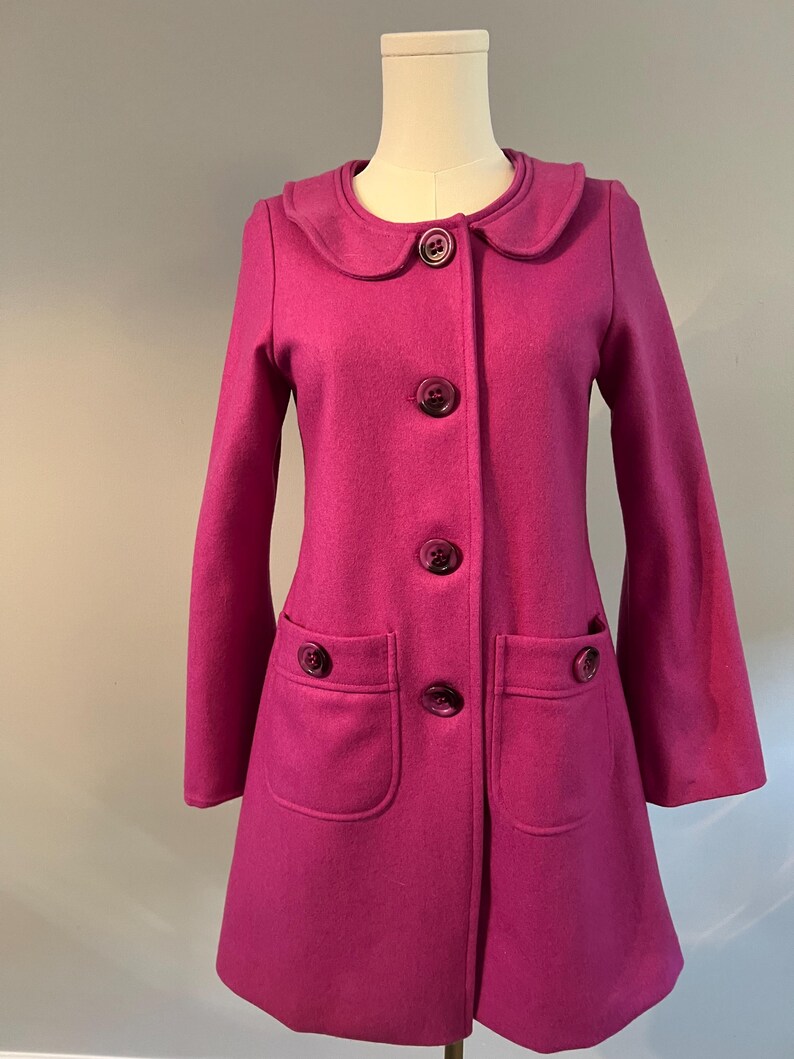 May include: A magenta coat with a rounded collar and large, matching buttons. The coat has two front pockets and an A-line shape. The coat is displayed on a mannequin.