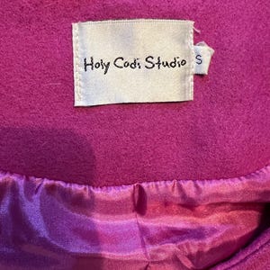 May include: A close-up of a magenta coat. The coat features a satin lining and a label that reads "Holy Codi Studio" with a size "S" tag. The coat's fabric appears to be a soft, felted material.
