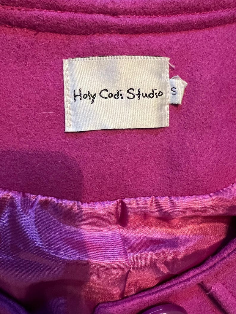 May include: A close-up of a magenta coat. The coat features a satin lining and a label that reads "Holy Codi Studio" with a size "S" tag. The coat's fabric appears to be a soft, felted material.