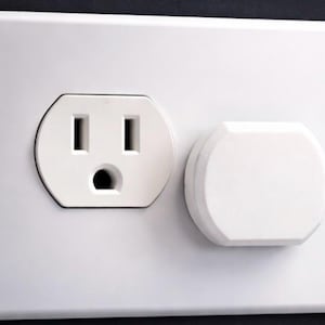 May include: A white electrical outlet with two vertical slots and a grounding hole, next to a white outlet cover. The outlet and cover are set in a white wall plate against a dark background.