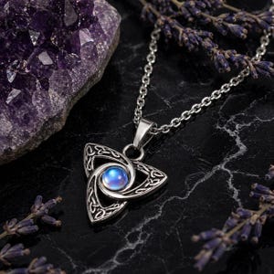 May include: Silver Celtic knot pendant necklace with a blue iridescent stone centre. The triangular pendant features detailed knotwork. The necklace is shown on a black surface with purple amethyst and lavender sprigs.