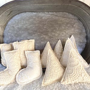 May include: A collection of small, cream-colored fabric ornaments featuring intricate lace patterns. The set includes several boot-shaped and triangular-shaped stuffed pieces with frayed edges, arranged in front of a large, oval, galvanized metal tub.