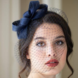 Può includere: Un fascinator blu navy caratterizzato da un fiocco strutturato e un delicato velo a gabbia che copre la parte superiore del viso. Il copricapo è realizzato in tessuto testurizzato ed è abbinato a un capo blu navy con decorazioni scure.