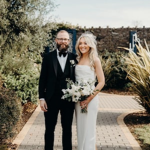 May include: A couple stands on a brick pathway, the woman in a white strapless gown and the man in a black tuxedo. The woman holds a bouquet of white flowers. The background includes greenery and a stone wall.