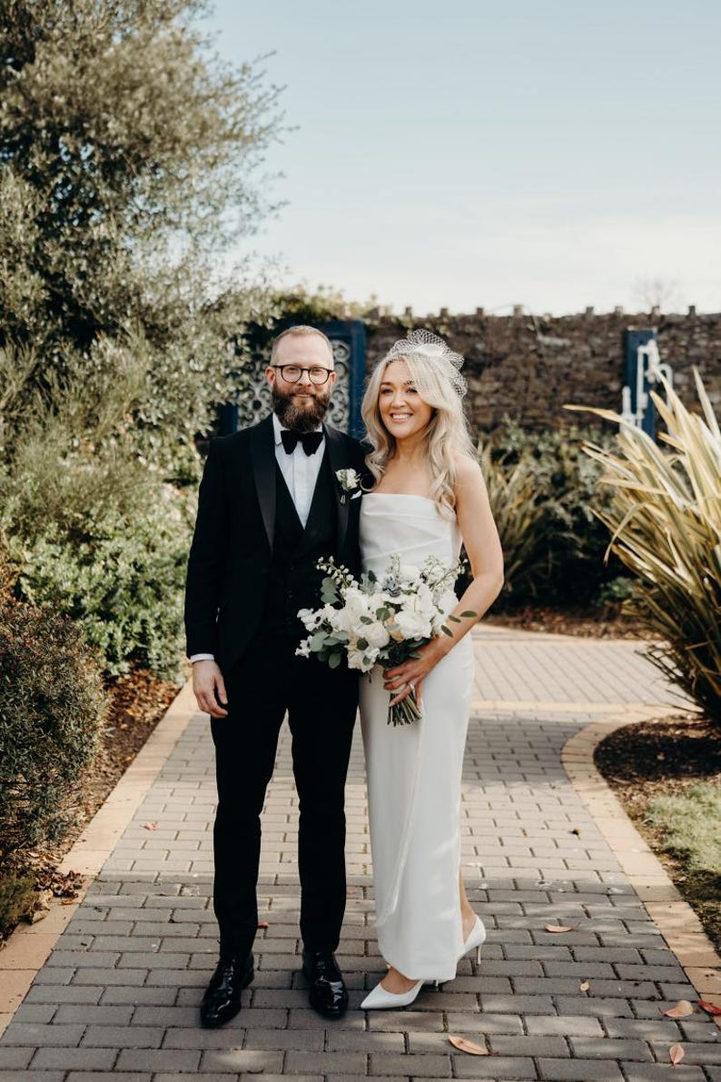 May include: A couple stands on a brick pathway, the woman in a white strapless gown and the man in a black tuxedo. The woman holds a bouquet of white flowers. The background includes greenery and a stone wall.