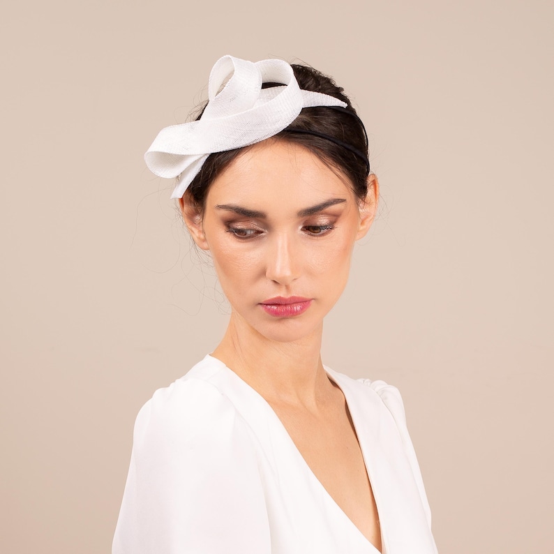 May include: A white fabric headband with a small, white, fabric flower accent. The headband is worn by a person with dark hair.