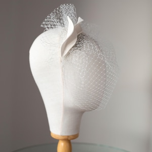 May include: A white headpiece with a veil, displayed on a mannequin head. The headpiece features a decorative accent and a delicate net veil. The mannequin is mounted on a wooden stand, set against a neutral background.
