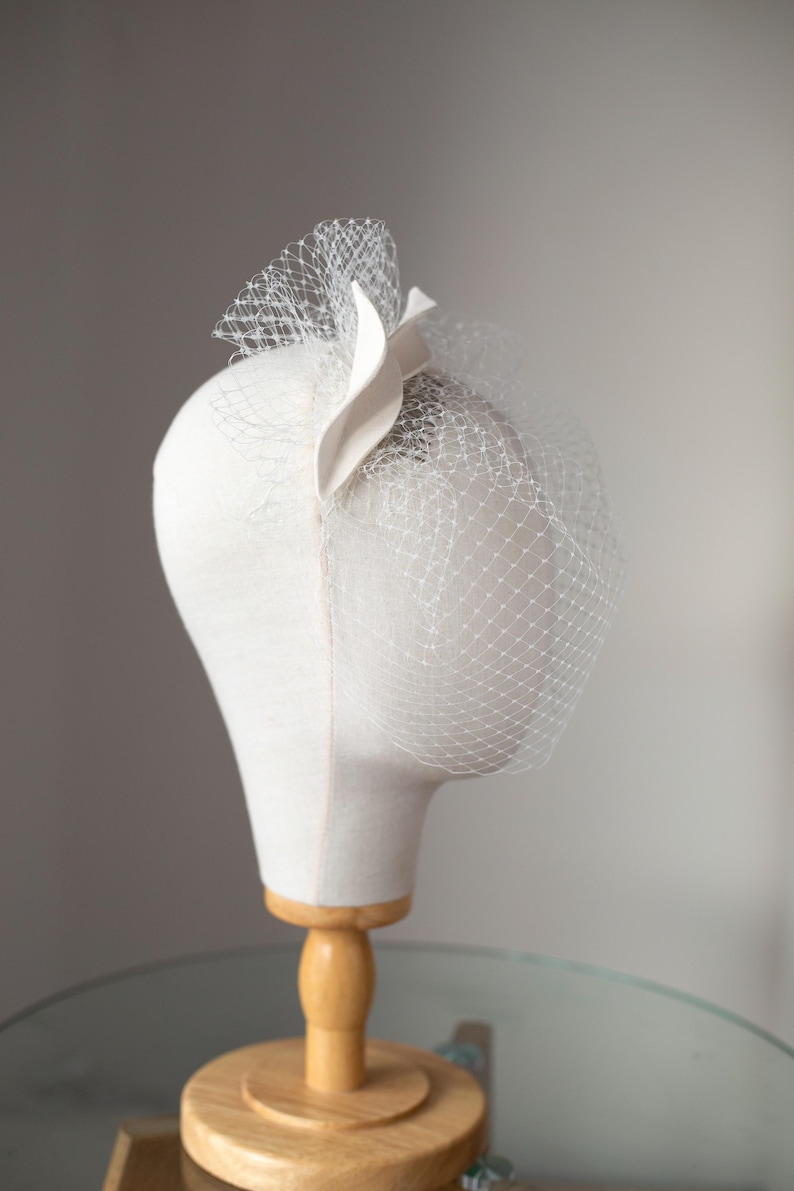 May include: A white headpiece with a veil, displayed on a mannequin head. The headpiece features a decorative accent and a delicate net veil. The mannequin is mounted on a wooden stand, set against a neutral background.