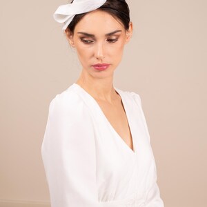May include: A white dress with a white fabric headband. The dress has a v-neck and long sleeves. The headband is tied in a bow on top of the head.