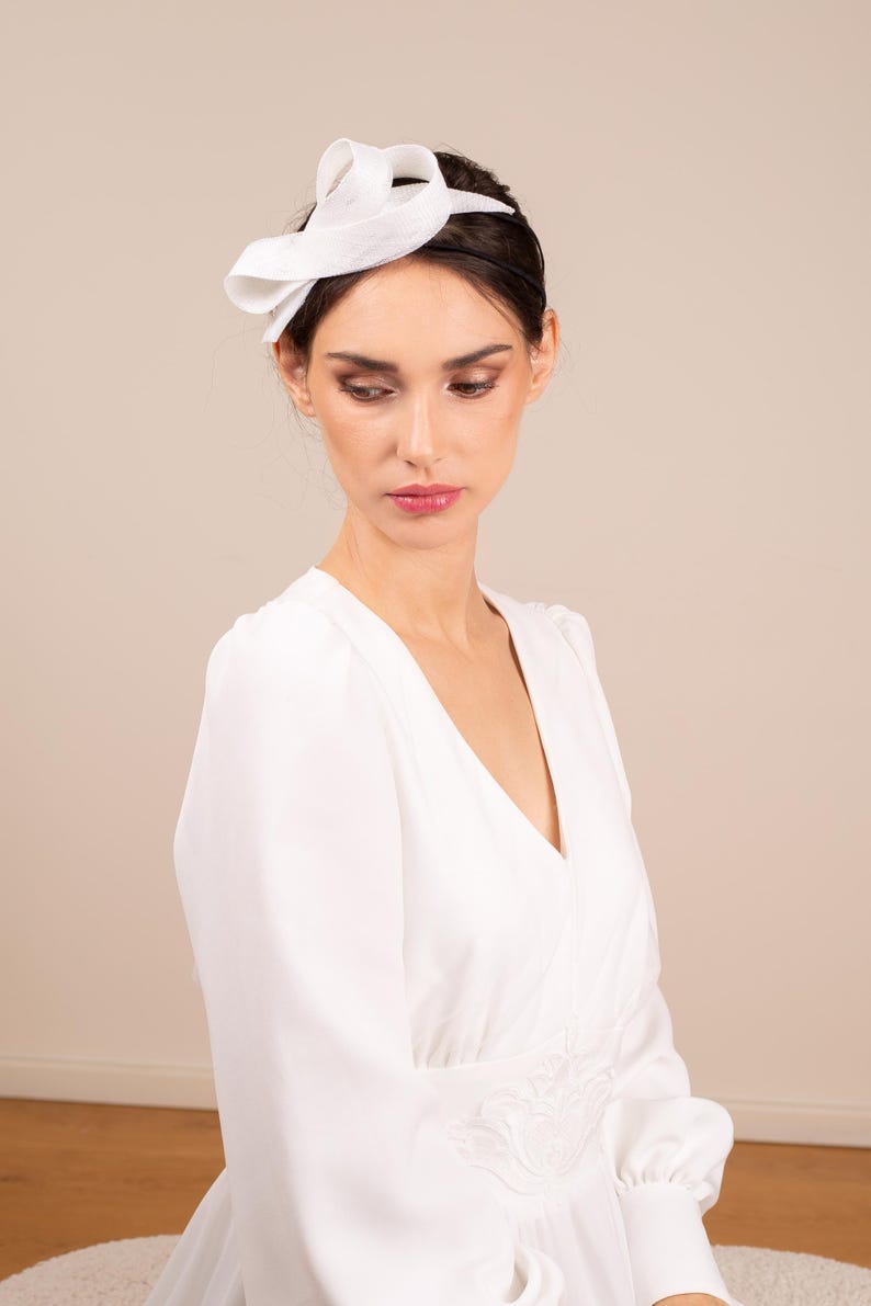 May include: A white dress with a white fabric headband. The dress has a v-neck and long sleeves. The headband is tied in a bow on top of the head.