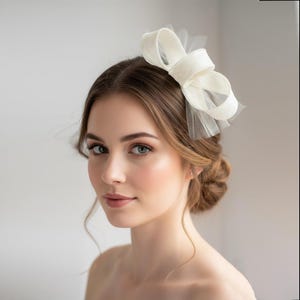 以下が含まれることがあります： オフホワイトのヘアアクセサリー。硬めの生地で作られたループ状の構造的なリボンが、繊細で透け感のあるチュールネットのベースに重ねられています。頭の横に配置されており、すっきりとしたレイヤードデザインが特徴です。