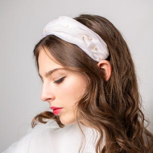 May include: A white satin headband adorned with small, faux pearls. The headband is wide and padded, creating a voluminous look. The pearls are scattered across the top, adding a touch of elegance. The headband is worn on a person with long, wavy brown hair.