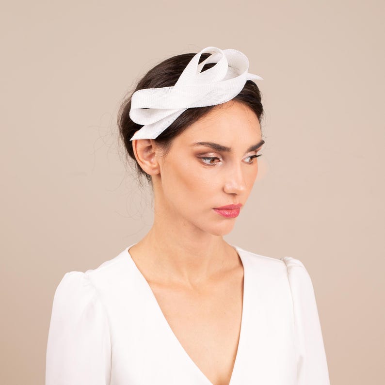 May include: A white fabric headband with a large bow on top. The headband is worn by a person with dark hair.