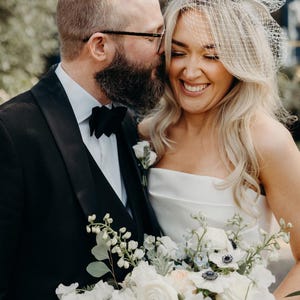 May include: A smiling bride and groom embrace, the groom in a black tuxedo with a bow tie, and the bride in a white strapless gown. She holds a bouquet of white flowers. The bride wears a veil and has blonde hair.