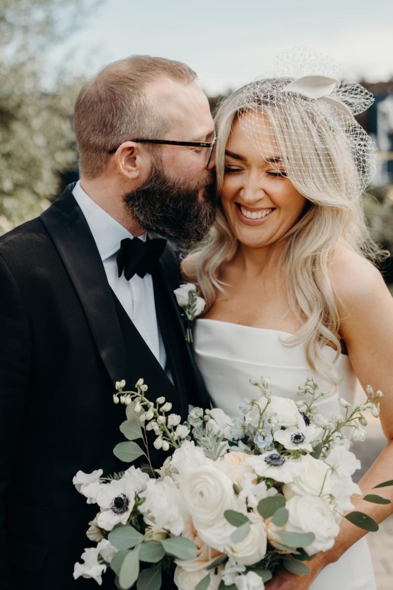 May include: A smiling bride and groom embrace, the groom in a black tuxedo with a bow tie, and the bride in a white strapless gown. She holds a bouquet of white flowers. The bride wears a veil and has blonde hair.