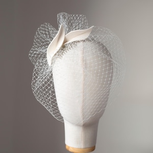 May include: A cream-colored headpiece with a delicate veil, displayed on a mannequin head. The headpiece features a fabric design with a net veil. The veil is a fine, open weave, and the headpiece is mounted on a wooden stand.