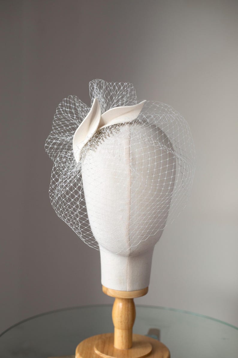 May include: A cream-colored headpiece with a delicate veil, displayed on a mannequin head. The headpiece features a fabric design with a net veil. The veil is a fine, open weave, and the headpiece is mounted on a wooden stand.