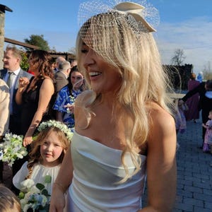May include: A woman in a white, strapless wedding dress smiles, surrounded by guests. She wears a veil headpiece. A young girl in a white dress and flower crown holds a bouquet. The scene is outdoors on a sunny day.
