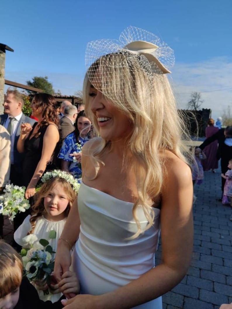 May include: A woman in a white, strapless wedding dress smiles, surrounded by guests. She wears a veil headpiece. A young girl in a white dress and flower crown holds a bouquet. The scene is outdoors on a sunny day.