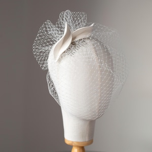 May include: A white headpiece with a veil, displayed on a mannequin head. The headpiece features a curved, petal-like accent and a delicate net veil. The mannequin is mounted on a wooden stand, set against a neutral background.