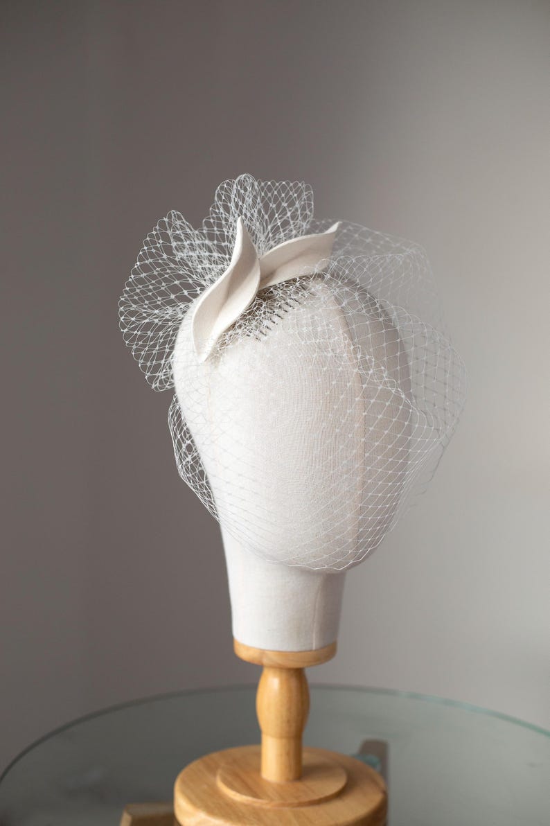 May include: A white headpiece with a veil, displayed on a mannequin head. The headpiece features a curved, petal-like accent and a delicate net veil. The mannequin is mounted on a wooden stand, set against a neutral background.