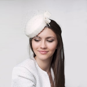 May include: A white felt pillbox hat with a white veil and a white bow. The hat is worn on the side of the head.