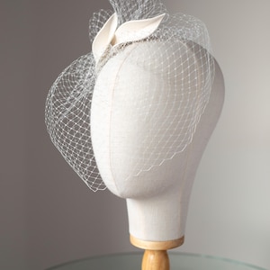 May include: A cream-colored headpiece with a delicate veil and decorative accents. The headpiece is displayed on a mannequin head with a wooden stand, showcasing its design and structure. The veil is a fine net, adding a touch of elegance.