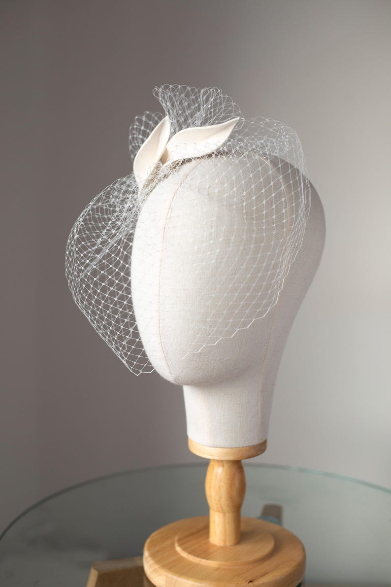 May include: A cream-colored headpiece with a delicate veil and decorative accents. The headpiece is displayed on a mannequin head with a wooden stand, showcasing its design and structure. The veil is a fine net, adding a touch of elegance.
