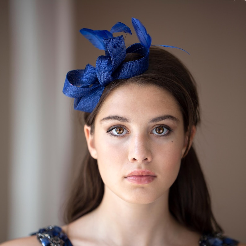 May include: A deep blue fascinator headpiece featuring a structured, looped fabric design accented with matching blue feathers. The accessory is positioned on the side of the head, showcasing a textured, woven material and vibrant blue tones.