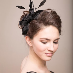 May include: A black fascinator headpiece featuring a textured, woven fabric bow design accented with several upright black feathers. The accessory is positioned on the side of the head, showcasing a structured, layered appearance with a dark finish.