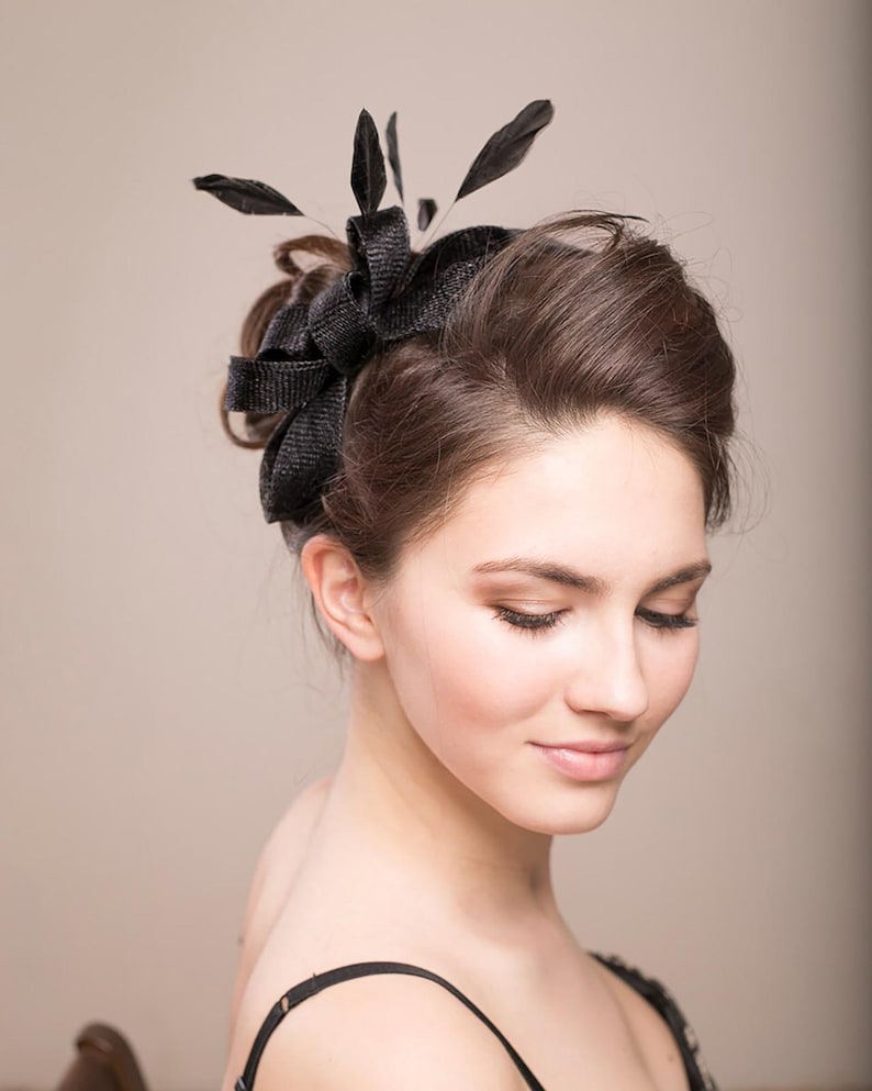 May include: A black fascinator headpiece featuring a textured, woven fabric bow design accented with several upright black feathers. The accessory is positioned on the side of the head, showcasing a structured, layered appearance with a dark finish.