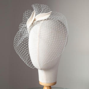 May include: A cream-colored fascinator with a delicate net veil, displayed on a mannequin head. The headpiece features a fabric leaf design and a fine mesh veil that drapes over the face. The mannequin is mounted on a wooden stand.