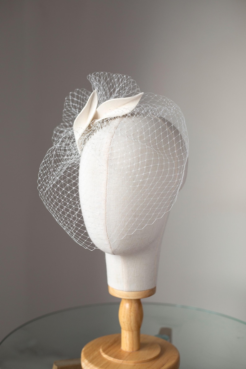 May include: A cream-colored fascinator with a delicate net veil, displayed on a mannequin head. The headpiece features a fabric leaf design and a fine mesh veil that drapes over the face. The mannequin is mounted on a wooden stand.