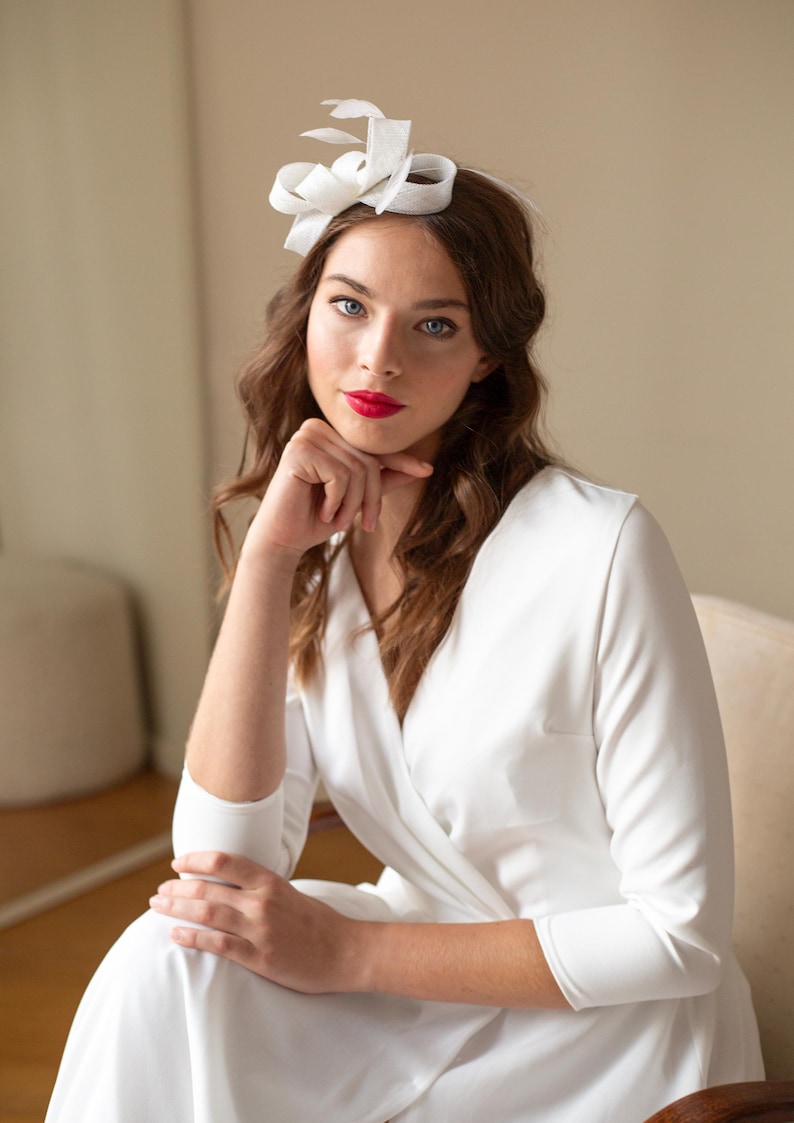 May include: A white fascinator headpiece featuring structured loops and delicate feather accents, worn as a hair accessory. The piece is crafted from a smooth, stiffened fabric, creating a sculptural, layered design that sits atop the hair.
