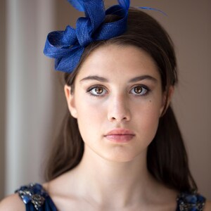 May include: A deep blue fascinator headpiece featuring a structured, woven loop design accented with matching blue feathers. The accessory is positioned on the side of the head, showcasing a textured fabric finish and vibrant blue color tones.