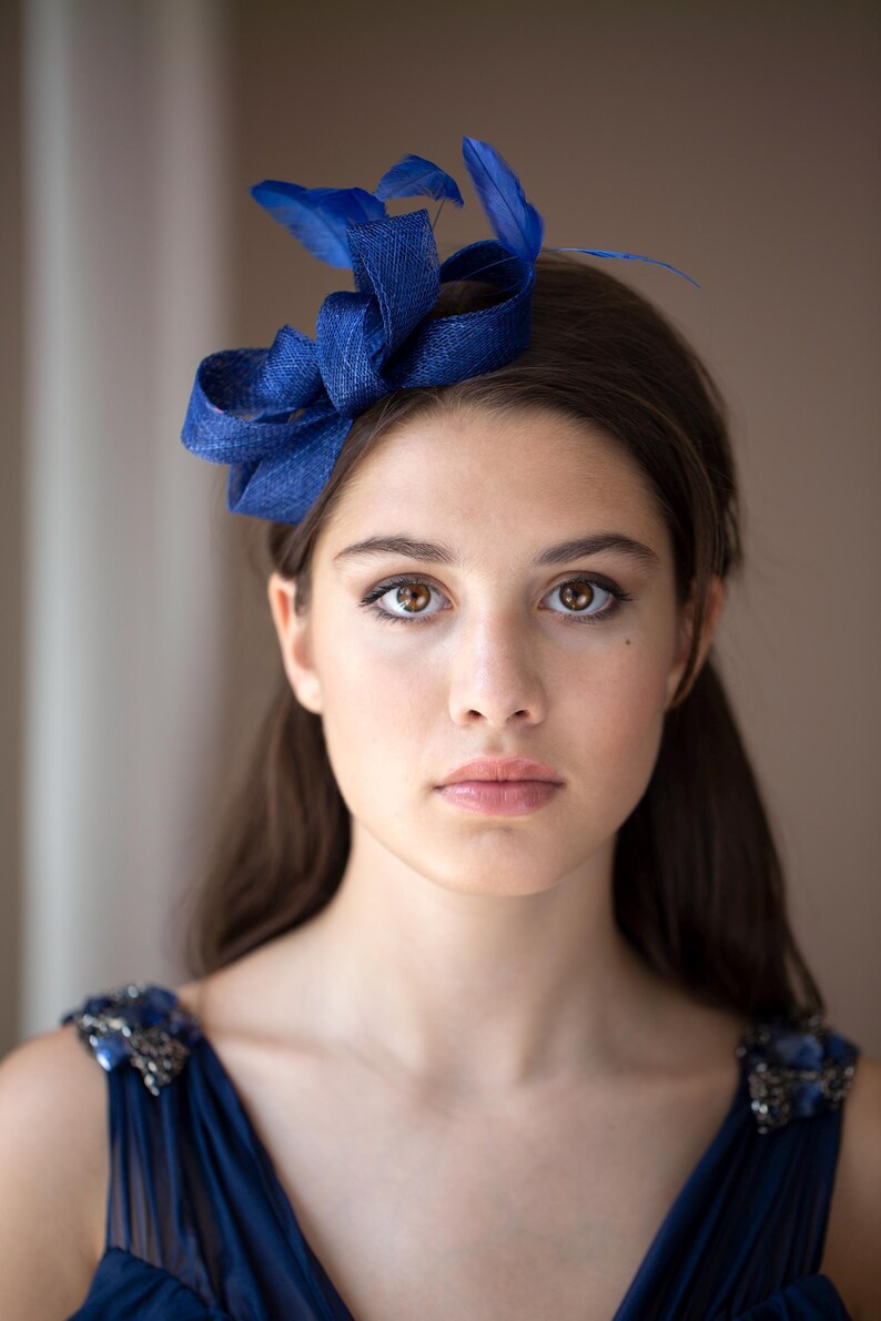 May include: A deep blue fascinator headpiece featuring a structured, woven loop design accented with matching blue feathers. The accessory is positioned on the side of the head, showcasing a textured fabric finish and vibrant blue color tones.
