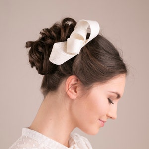 以下が含まれることがあります： 女性向けの白い布製のヘアアクセサリー。リボンは大きく緩く結ばれており、柔らかく質感のある布で作られています。