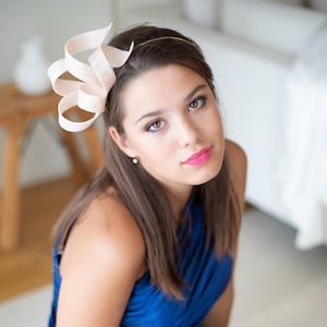 Puede incluir: Tocado rosa claro con un diseño en espiral, sujeto a una fina diadema dorada. El tocado lo lleva una persona que viste un vestido azul. La persona tiene el pelo largo castaño y mira a la cámara.