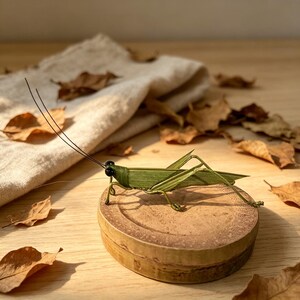 Puede incluir: Una escultura artesanal de saltamontes hecha de hojas verdes y alambre, con ojos de cuentas negras y largas antenas. El insecto está posado sobre una pequeña base redonda de madera. El fondo incluye hojas secas y un paño beige.