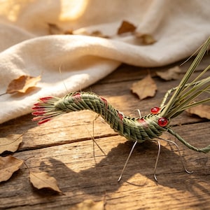 May include: A handcrafted decorative insect sculpture made of woven green and red plant material. The insect has red bead eyes and wire legs, resting on a wooden surface with fallen leaves. The sculpture is a unique piece of home decor.